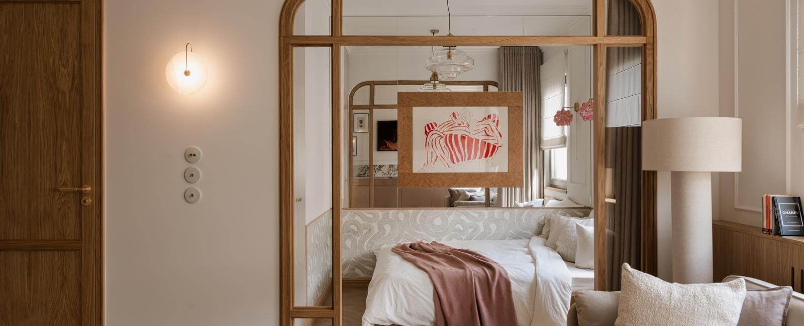 Nowoczesny pokój hotelowy z łóżkiem i miękką pościelą, drewnianą ramą lustra, lampą stołową i przytulną sofą z poduszkami.