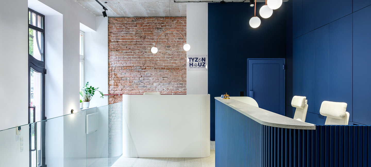 Nowoczesna recepcja hotelowa z granatowym biurkiem, białą ladą do zameldowania, odsłoniętą cegłą, kulistymi lampami i szklaną balustradą.