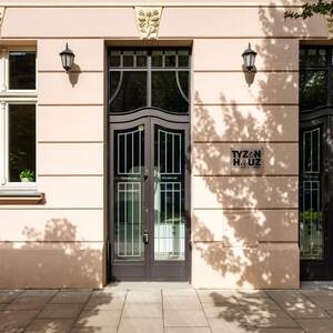 Elegancki wejście do hotelu z ozdobnymi drzwiami i kremową fasadą, z zewnętrznymi lampami i roślinnymi dekoracjami.