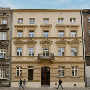 Odrestaurowana fasada hotelu z ozdobnymi nadokiennymi detalami, żeliwnym balkonem i wejściem od ulicy, przechodnie.