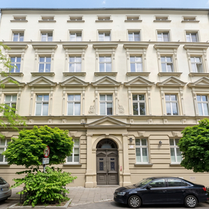 Elegancka kremowa elewacja kamienicy z ozdobnymi nadokiennikami i łukowym wejściem, ulica z drzewami i zaparkowanymi samochodami.