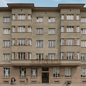 Symetryczna elewacja kamienicy z dekoracyjnymi balkonami, zdobionymi panelami okiennymi, sklepami i zaparkowanym rowerem.