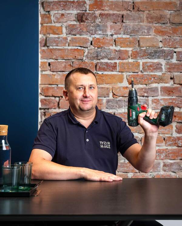 Uśmiechnięty konserwator trzyma wiertarkę akumulatorową przed odsłoniętą ceglaną ścianą, stół z karafką wody i szklankami