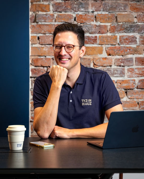 Uśmiechnięty mężczyzna w koszulce z logo Tyzen Hauz przy biurku z laptopem i kawą na tle ceglanej ściany.