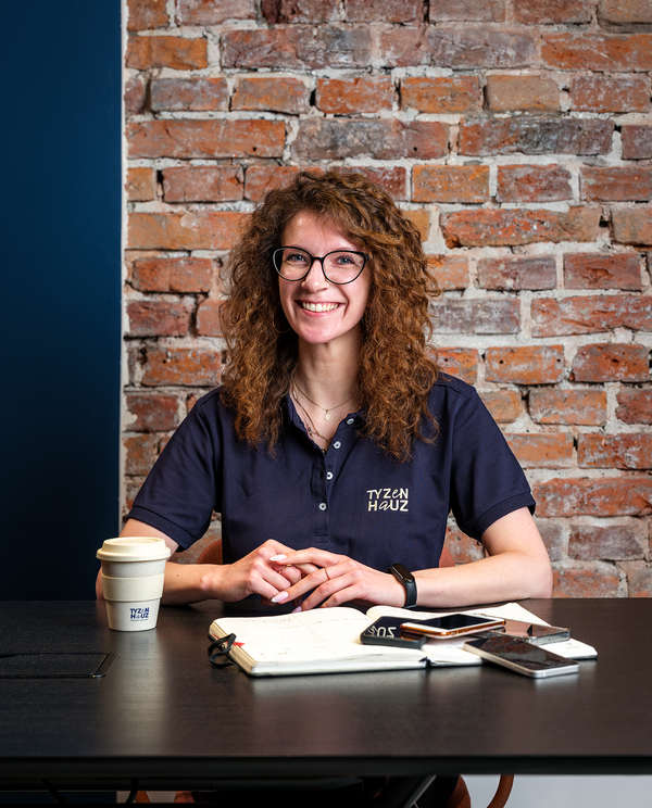 Recepcjonistka hotelowa w granatowej koszulce z logo Tyzen Hauz siedzi przy biurku z notatnikiem, telefonami i kawą na wynos