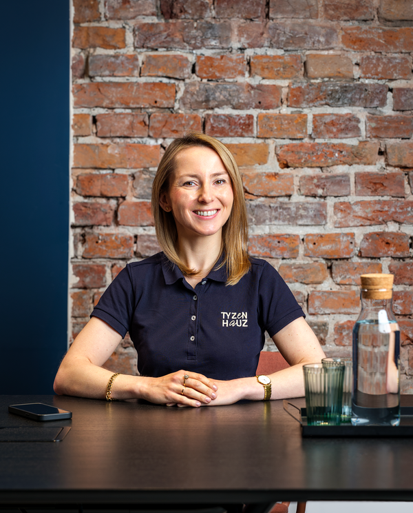 Recepcjonistka w koszulce z logo hotelu Tyzen Hauz siedzi przy nowoczesnym biurku na tle ceglanej ściany