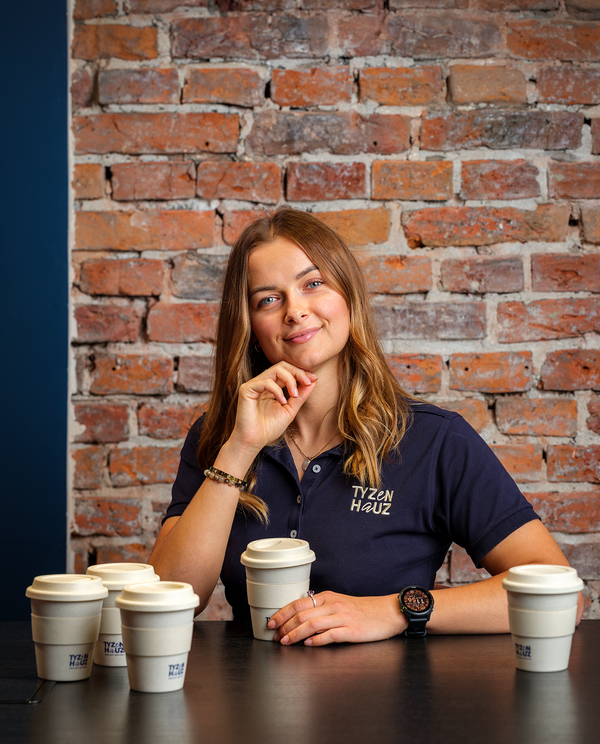 Uśmiechnięta kobieta za czarnym stołem z pięcioma kubkami na wynos w kawiarni Tyzen Hauz przy ścianie z czerwonej cegły