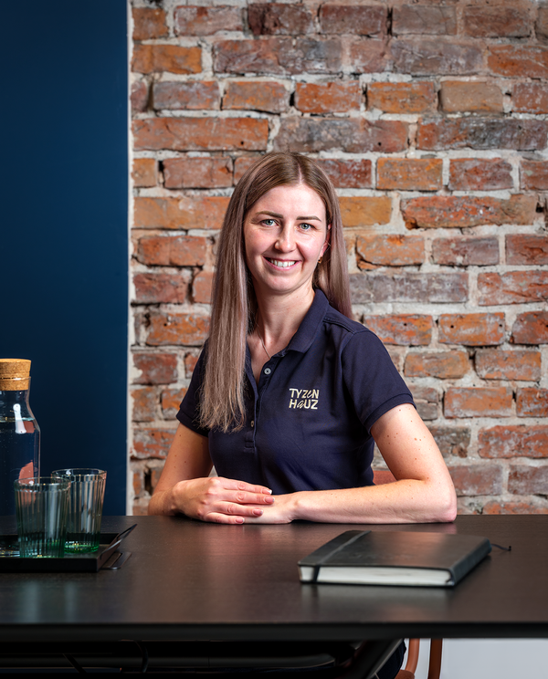 Uśmiechnięta kobieta w koszulce z logo hotelu siedzi przy czarnym stole, za nią surowa ceglana ściana, na stole dzbanek z wodą i zeszyt.