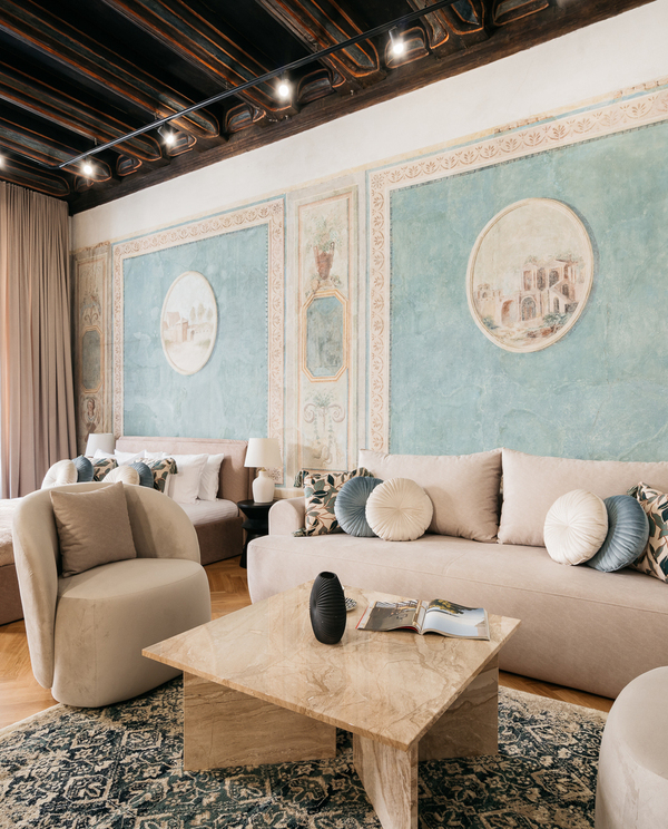 Historic suite with painted fresco walls, dark wood‑beamed ceiling, large window, plush beige sofa and bed, marble coffee table.