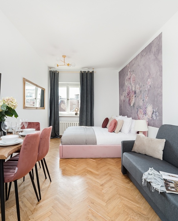 Bright studio suite with double bed by floral mural, set dining table, pink velvet chairs, wall TV and grey sofa.