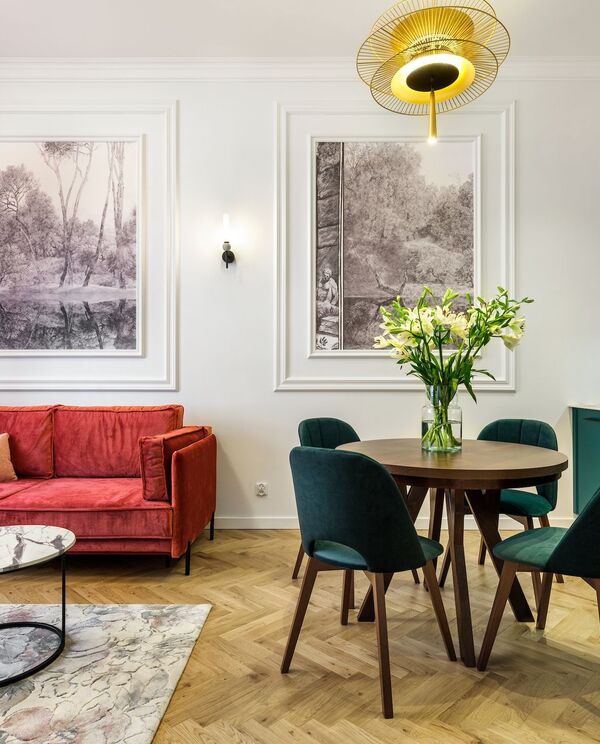 Przytulny apartament z czerwoną welurową sofą, okrągłym stołem i zielonymi krzesłami, wazon z liliami, turkusowy aneks i złota lampa.