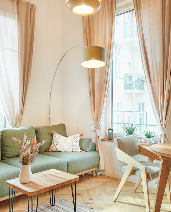 Jasny, przytulny salon w apartamencie z zieloną sofą, łukową lampą, drewnianym stolikiem i dużymi oknami z beżowymi zasłonami.
