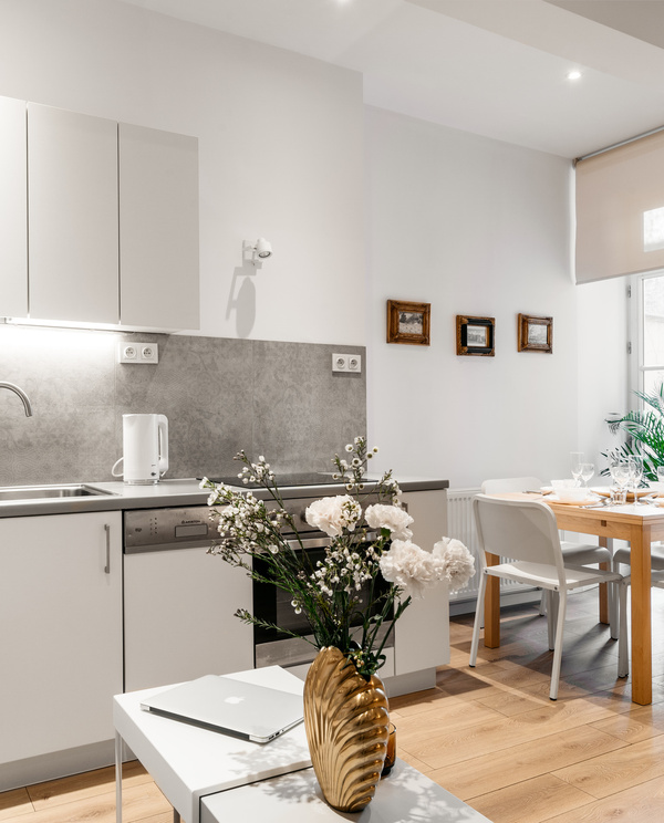 Bright modern kitchenette with white cabinets, stainless sink, electric kettle and a sunlit dining table by the window.