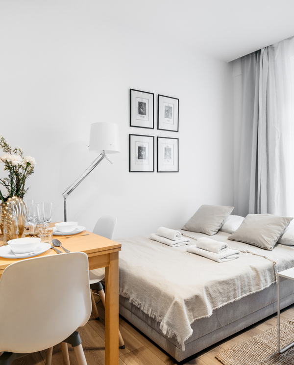 Bright minimalist studio with comfortable double bed, folded towels, dining table set for four, sheer curtains, lamp and decorative vase.