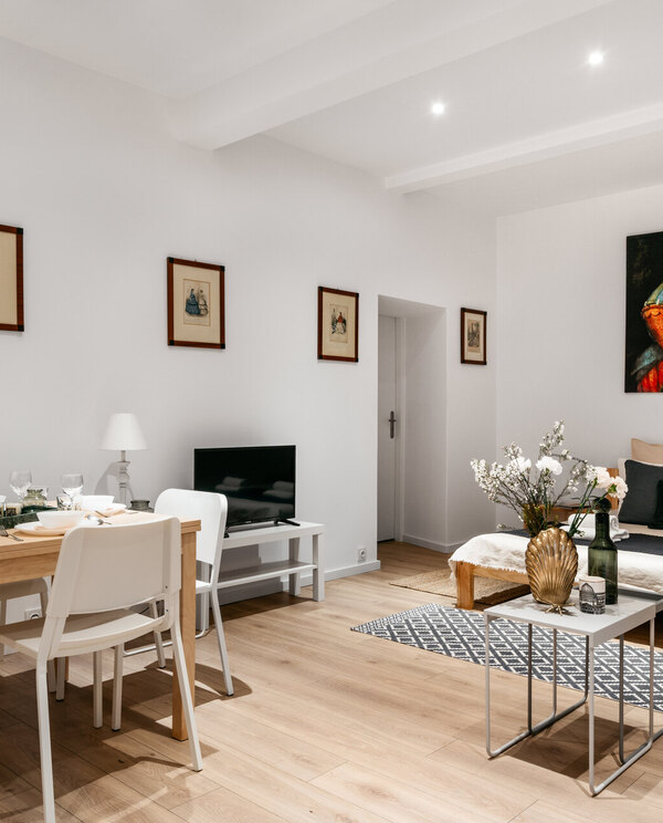 Bright studio suite with dining table set, queen bed with layered pillows, art, TV, vase of flowers, hardwood floors