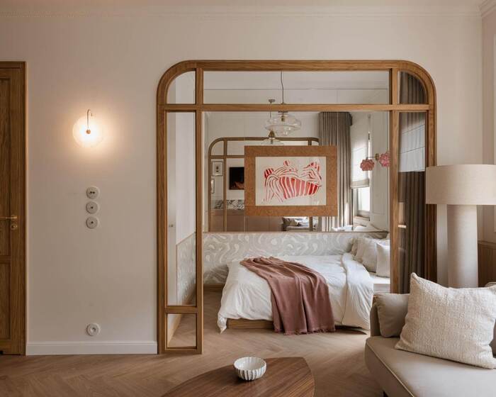 Modern hotel room with bed and soft bedding, wooden mirror frame, table lamp and cozy sofa with pillows.