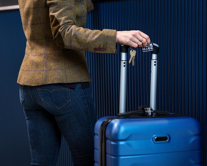 Gość ubrany w casualową marynarkę i dżinsy trzyma klucze oraz identyfikator przy uchwycie niebieskiej walizki podróżnej