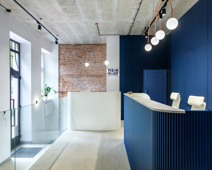Nowoczesna recepcja hotelowa z granatowym biurkiem, białą ladą do zameldowania, odsłoniętą cegłą, kulistymi lampami i szklaną balustradą.