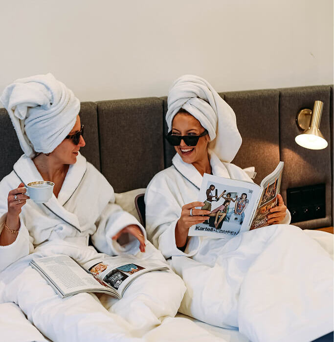Dwie kobiety w hotelowym łóżku, ubrane w białe szlafroki i turbany, relaksują się z kawą i czasopismami.