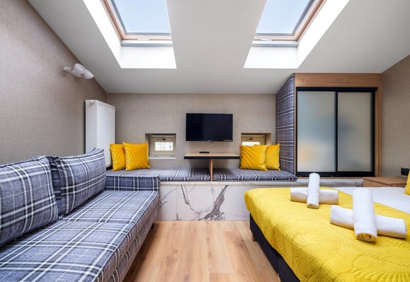 Modern hotel room with bed in yellow bedspread and comfortable plaid sofa under two large skylights.