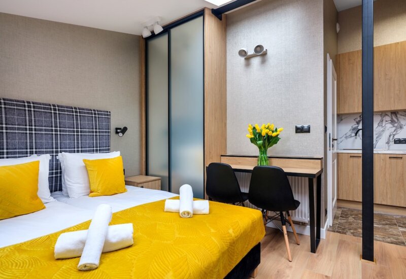 Modern hotel room with double bed, yellow accents, desk with two chairs and kitchen in the background.