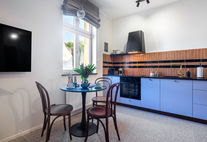 Hotel kitchen with light blue cabinets, black countertop and modern oven, at round table with flowers and cups.