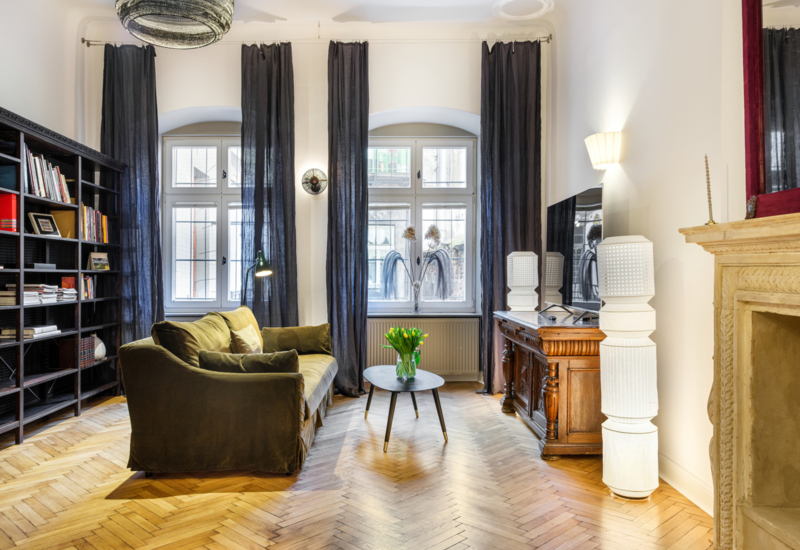 Stylish lounge with olive sofa, wooden floor, tall windows and elegant bookshelf.