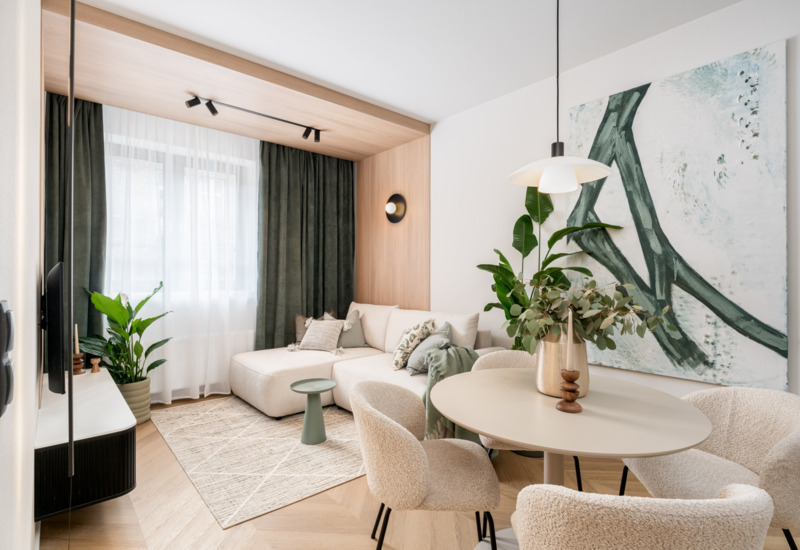 Modern hotel lounge with beige corner sofa, round table and soft armchairs accompanied by plants and large painting.