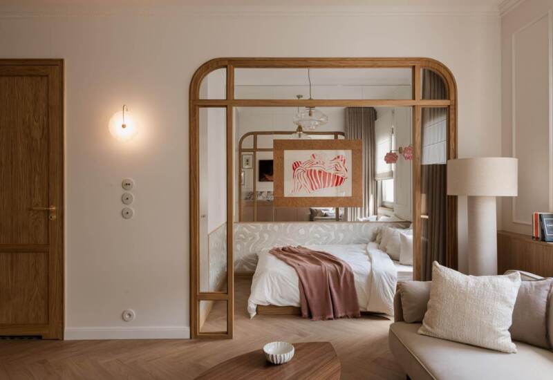 Modern hotel room with bed and soft bedding, wooden mirror frame, table lamp and cozy sofa with pillows.
