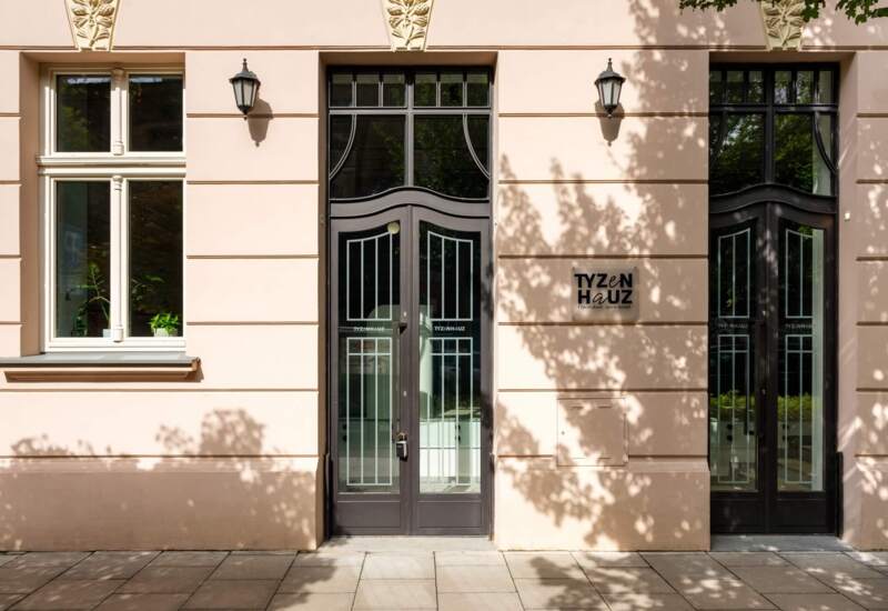 Elegant hotel entrance with decorative doors and cream facade, with exterior lamps and plant decorations.