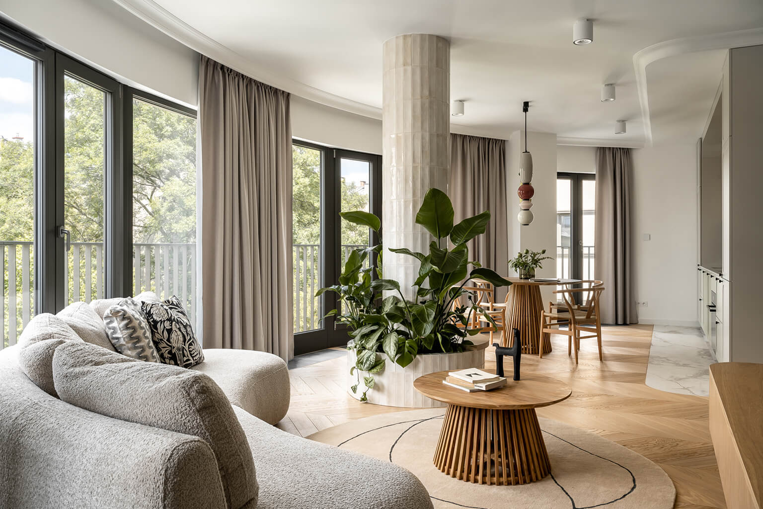 Nowoczesny apartament z dużymi oknami, jasnymi zasłonami, elegancką sofą, wewnętrzną rośliną i drewnianym stołem jadalnym.