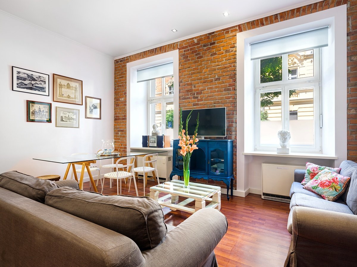 Przytulny salon z ceglaną ścianą, dwoma szarobrązowymi sofami, szklanym stołem i dużymi oknami zapewniającymi naturalne światło.