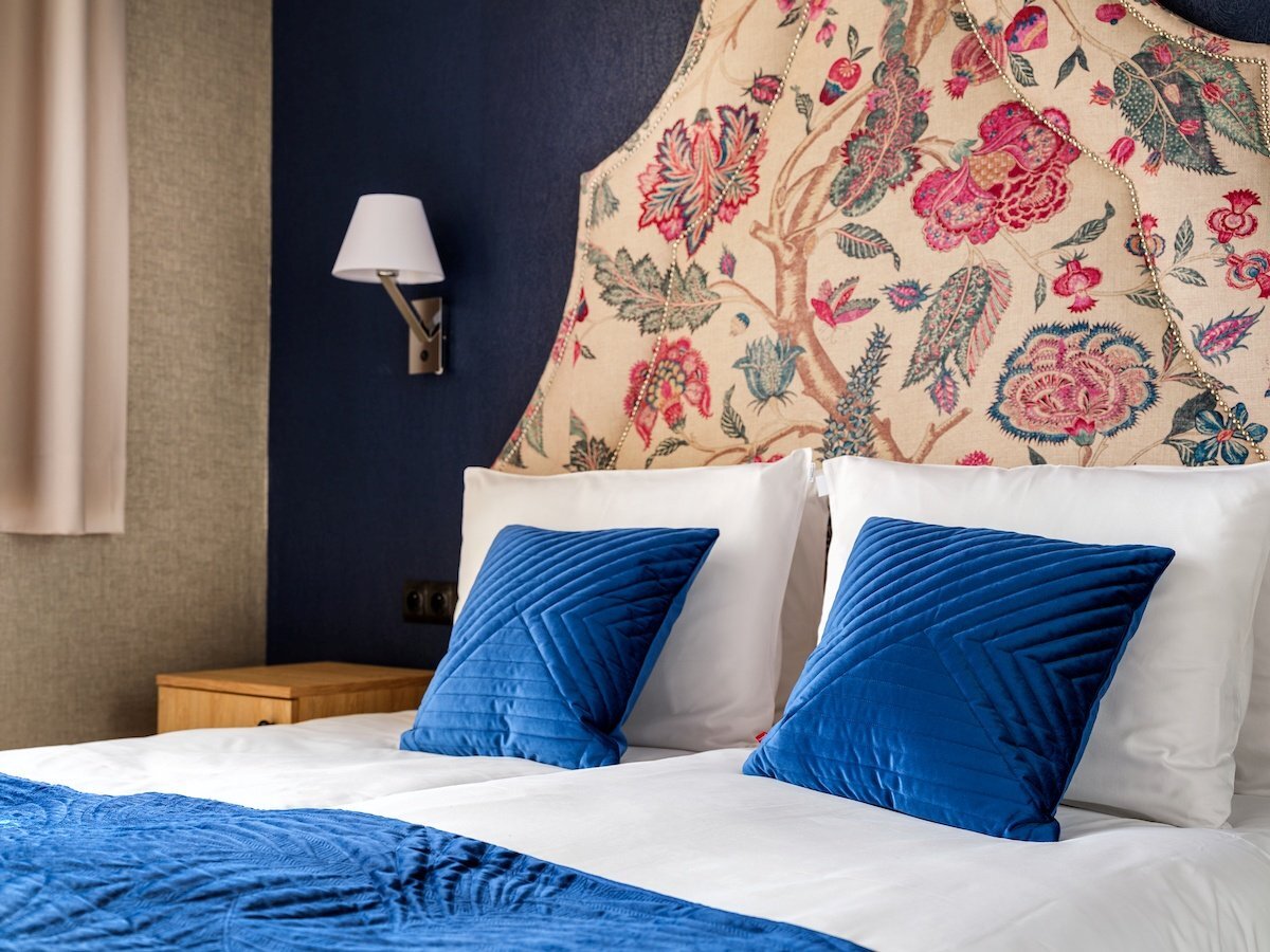 Dwa pojedyncze łóżka z białą pościelą, niebieskimi aksamitnymi poduszkami i narzutą, kwiatowe wezgłowie, kinkiet i drewniany stolik nocny.