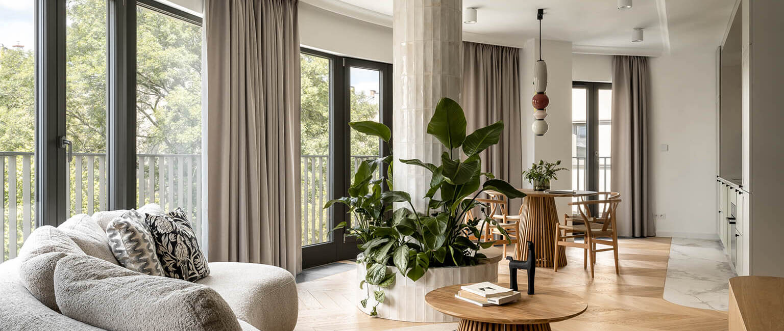Nowoczesny apartament z dużymi oknami, jasnymi zasłonami, elegancką sofą, wewnętrzną rośliną i drewnianym stołem jadalnym.