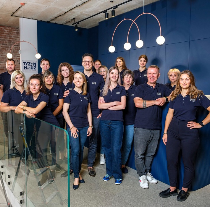 Zespół hotelu w jednakowych koszulkach, uśmiechnięci na nowoczesnej recepcji z niebieską ścianą, łukowymi lampami, cegłą i szklaną balustradą