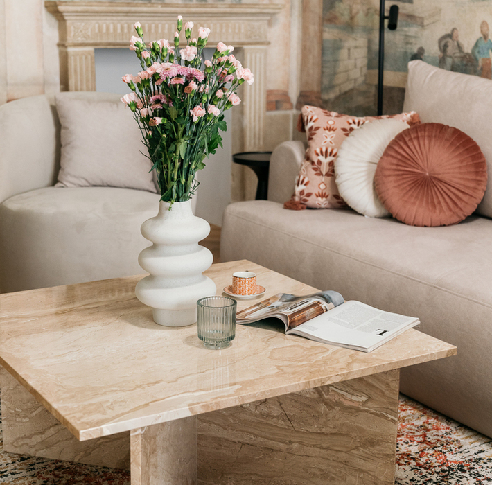 Butikowy salon z marmurowym stolikiem, wazonem z różowymi kwiatami, jasną sofą, ozdobnymi poduszkami i muralem nad kominkiem.