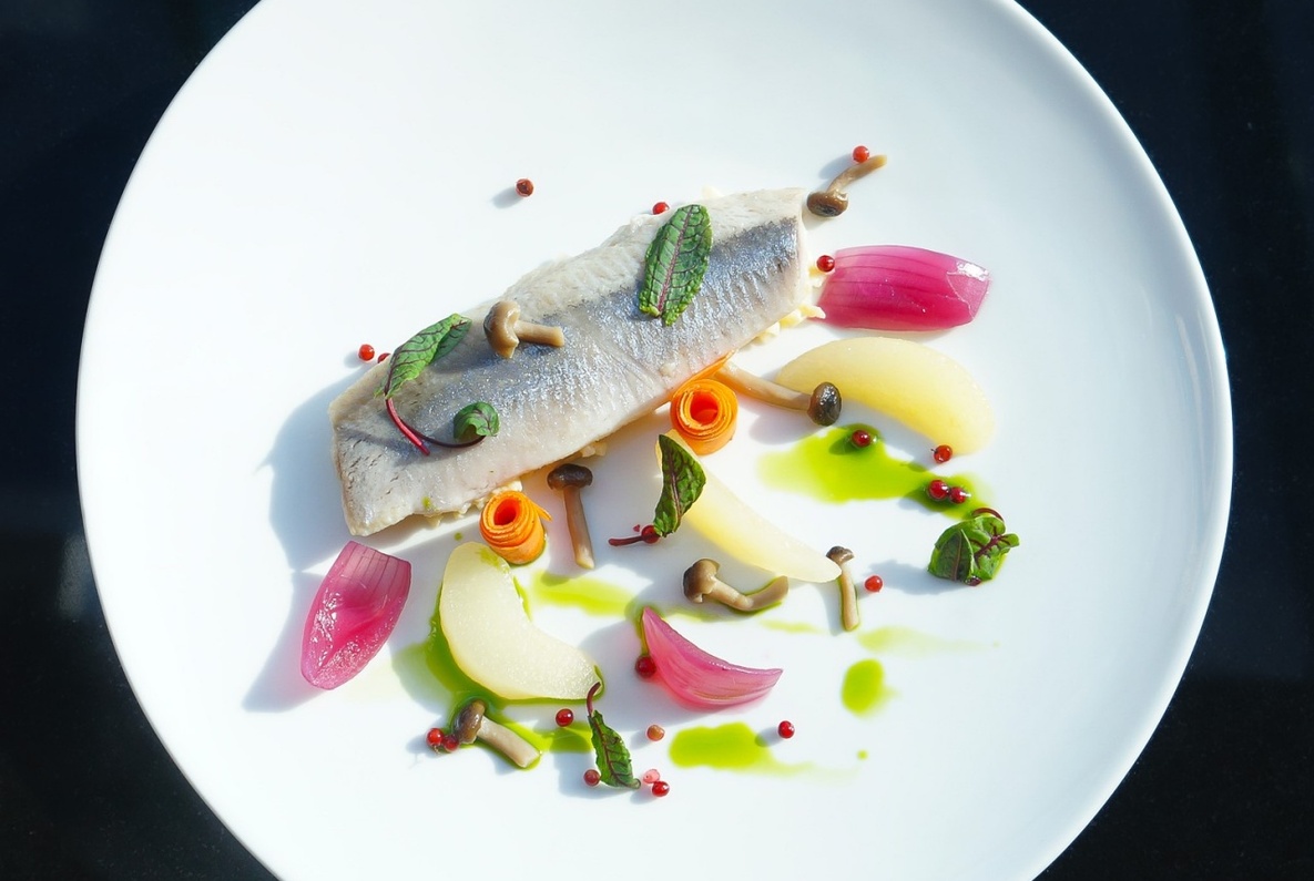 Delicate white fish fillet with pear slices, pickled red onions, carrot curls, mushrooms, pink peppercorns and herb oil on a white plate