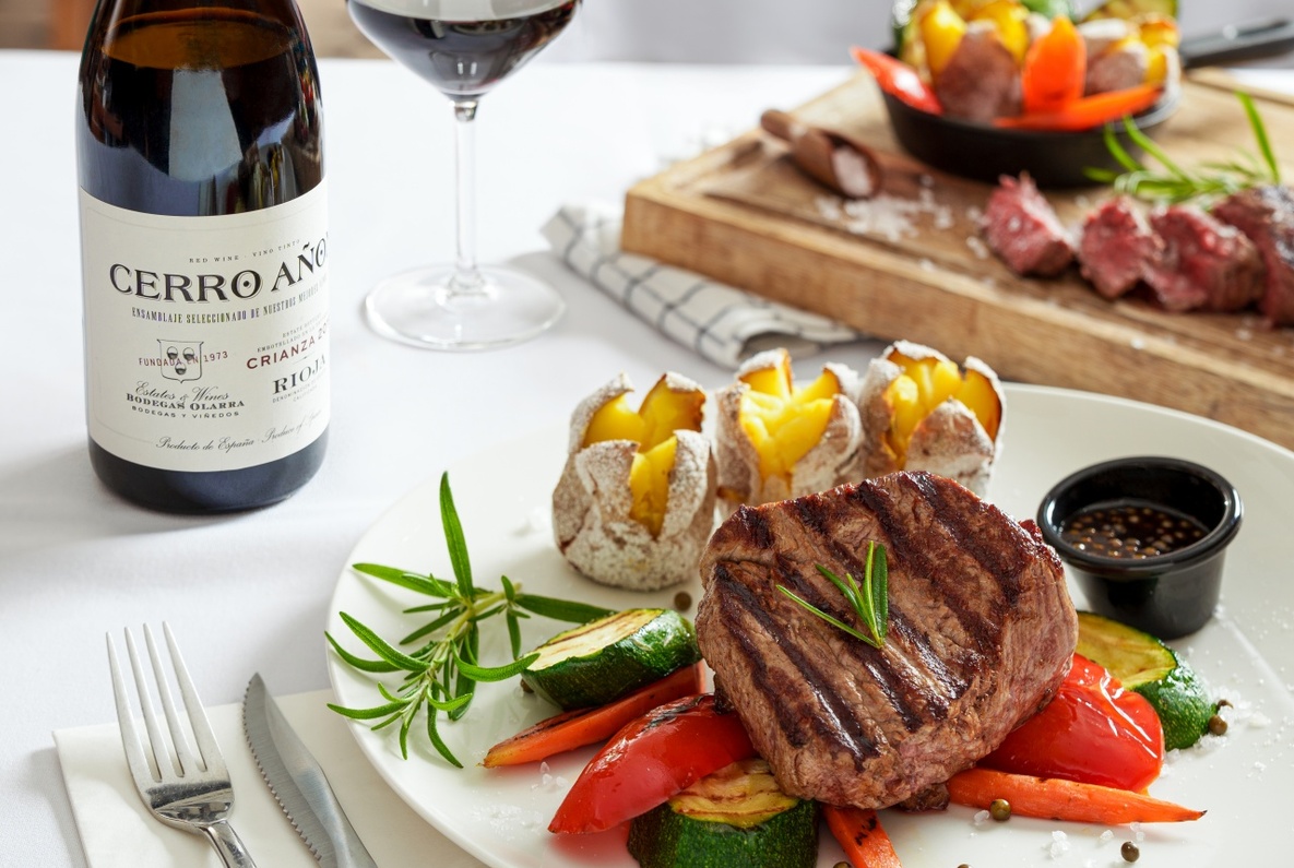 Grilled steak with roasted zucchini, peppers and baked baby potatoes, rosemary sprig and sauce, bottle and glass of red wine on white tablecloth