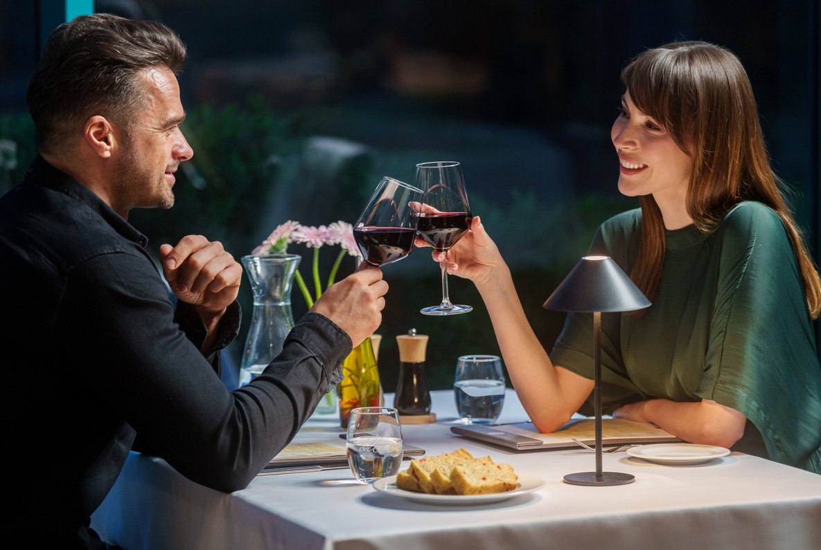 Couple toasting with red wine at a softly lit hotel restaurant table with bread, water, flowers and a modern table lamp