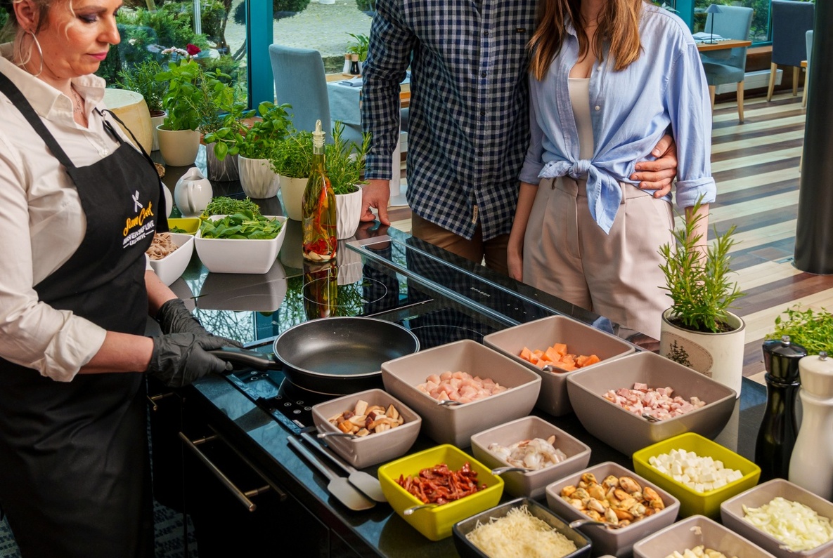 Couple watching chef prepare fresh ingredients at hotel live-cooking station in bright glass-walled dining area