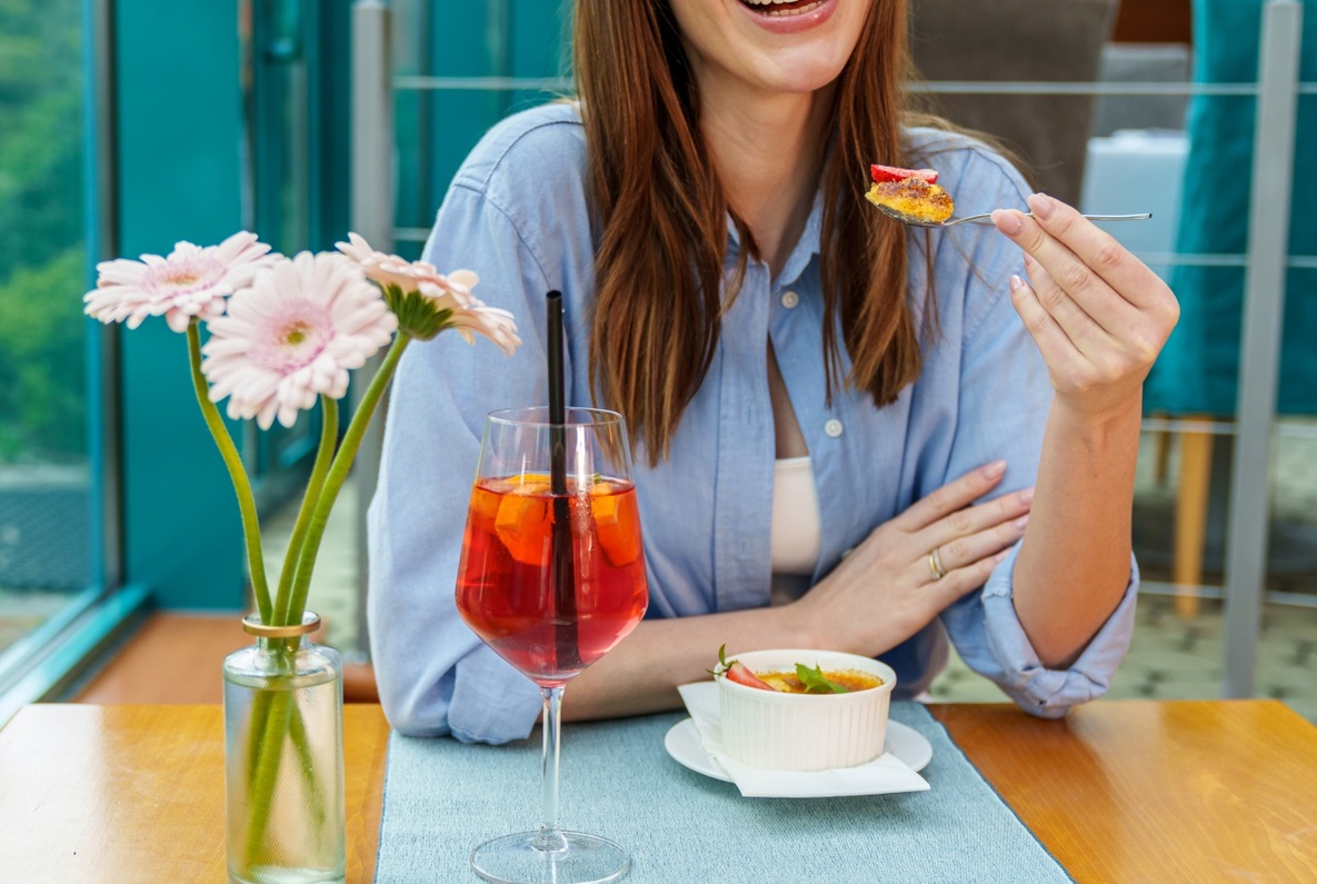 Smiling woman enjoying dessert and a red spritz at a glass-roofed hotel dining area with fresh flowers.