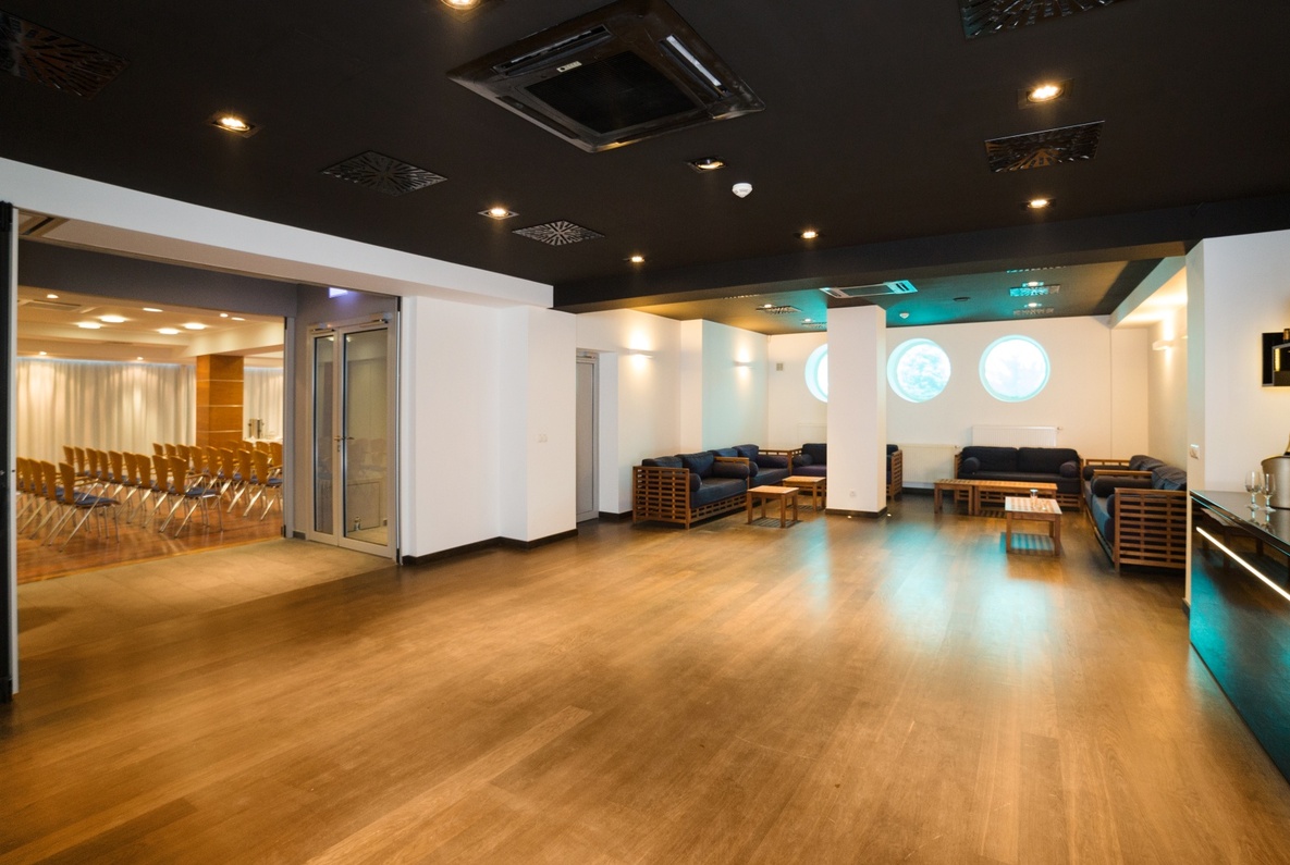 Spacious hotel event lounge with wooden floor, folding partition to conference room, navy sofas and round windows.