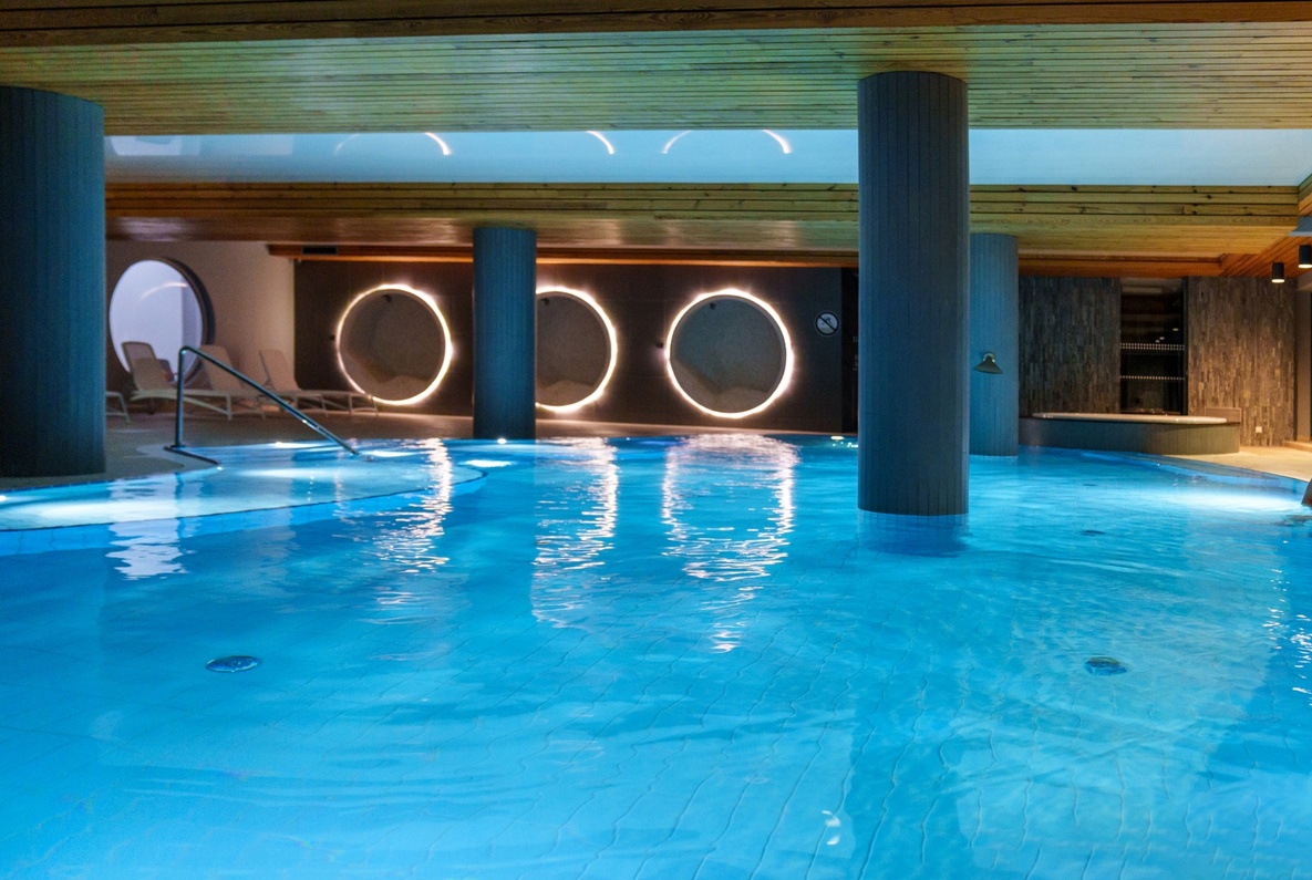 Kryty hotelowy basen spa z okrągłymi podświetlonymi panelami, drewnianym sufitem, leżakami i parą przy krawędzi.