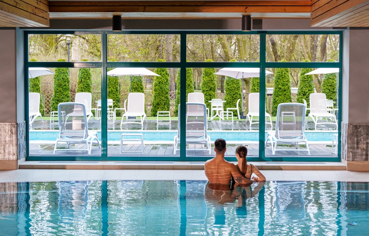 Couple relaxing in indoor hotel pool, sitting arm-in-arm facing large windows with sun loungers, umbrellas and garden view