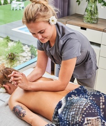 Terapeutka wykonuje relaksacyjny masaż pleców w jasnym gabinecie spa z dużym oknem, widokiem na ogród i basen