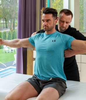 Terapeuta pomaga gościowi w rozciąganiu barków w gabinecie spa z widokiem na ogród i basen hotelowy.