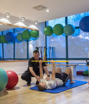 Personal trainer assisting guest with barbell press on mat in hotel fitness studio with exercise balls and kettlebells