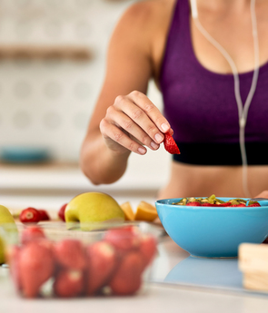 Fit guest in sports bra adding a strawberry to a blue breakfast bowl with fresh apples, strawberries and a blender jar