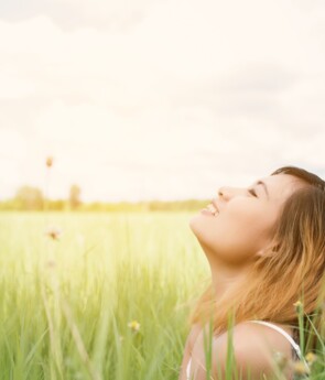 Woman sitting in tall green grass, smiling with her face lifted toward the sun in a bright, peaceful meadow