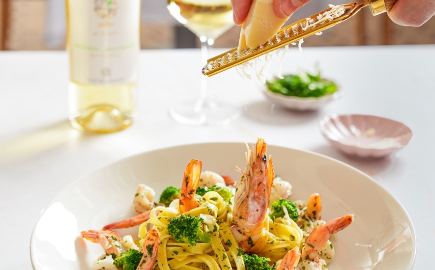 Tagliatelle z krewetkami i brokułem, doprawiane świeżo startym parmezanem przy butelce i kieliszku białego wina.
